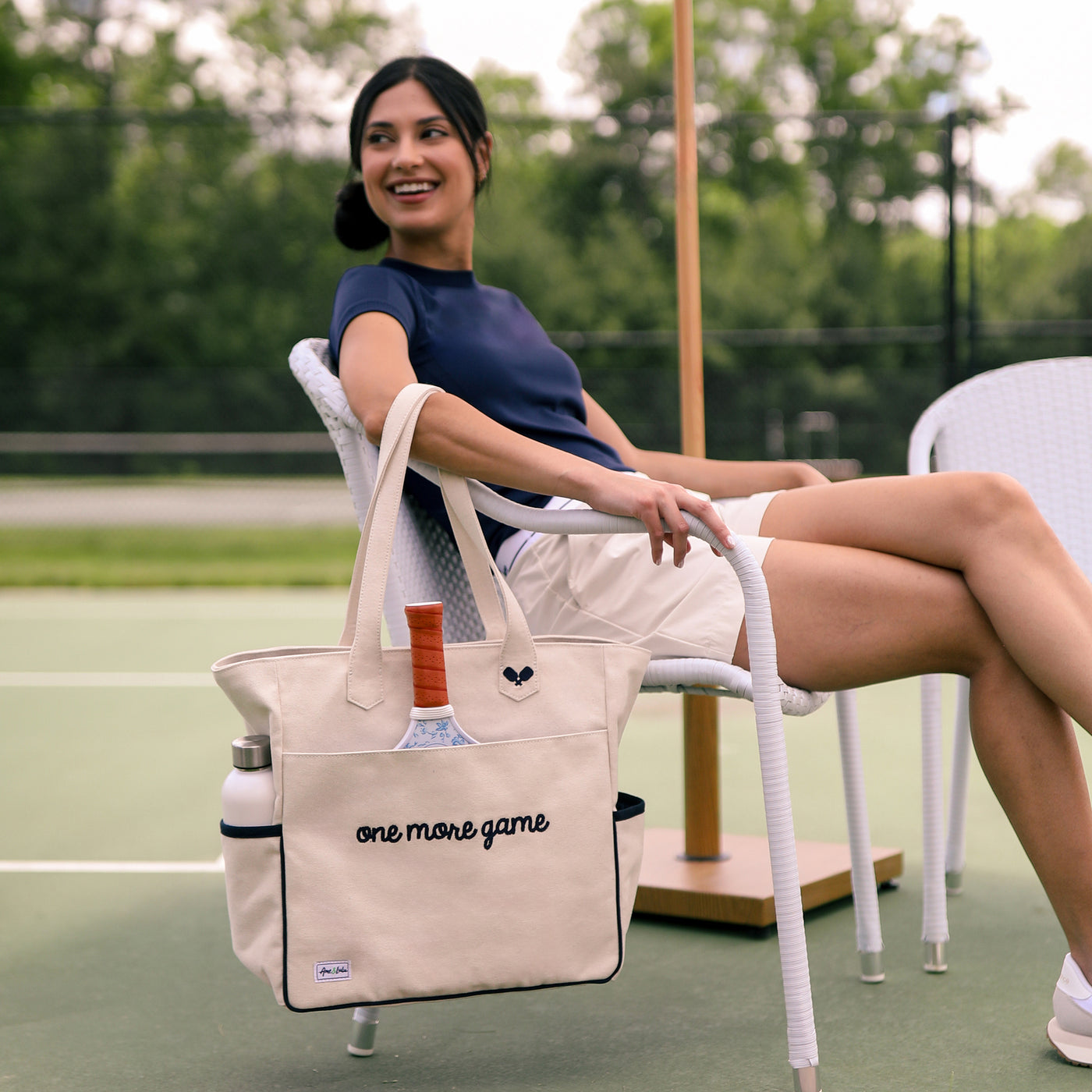 Tan canvas pickleball tote with slip pocket on front with a pickleball paddle inside. Front has navy embroidery that reads "one more game".