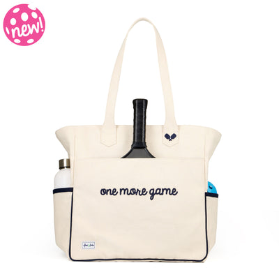 Tan canvas pickleball tote with slip pocket on front with a pickleball paddle inside. Front has navy embroidery that reads "one more game".