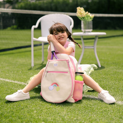 Natural canvas kids tennis backpack with pastel pink trim and water bottle pockets. There is a front tennis racquet pocket. A rainbow with a tennis ball is embroidered on the front pocket. 