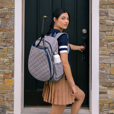 Women's tennis backpack with quilted grey nylon fabric and navy trim. The front pocket is holding a tennis racquet.