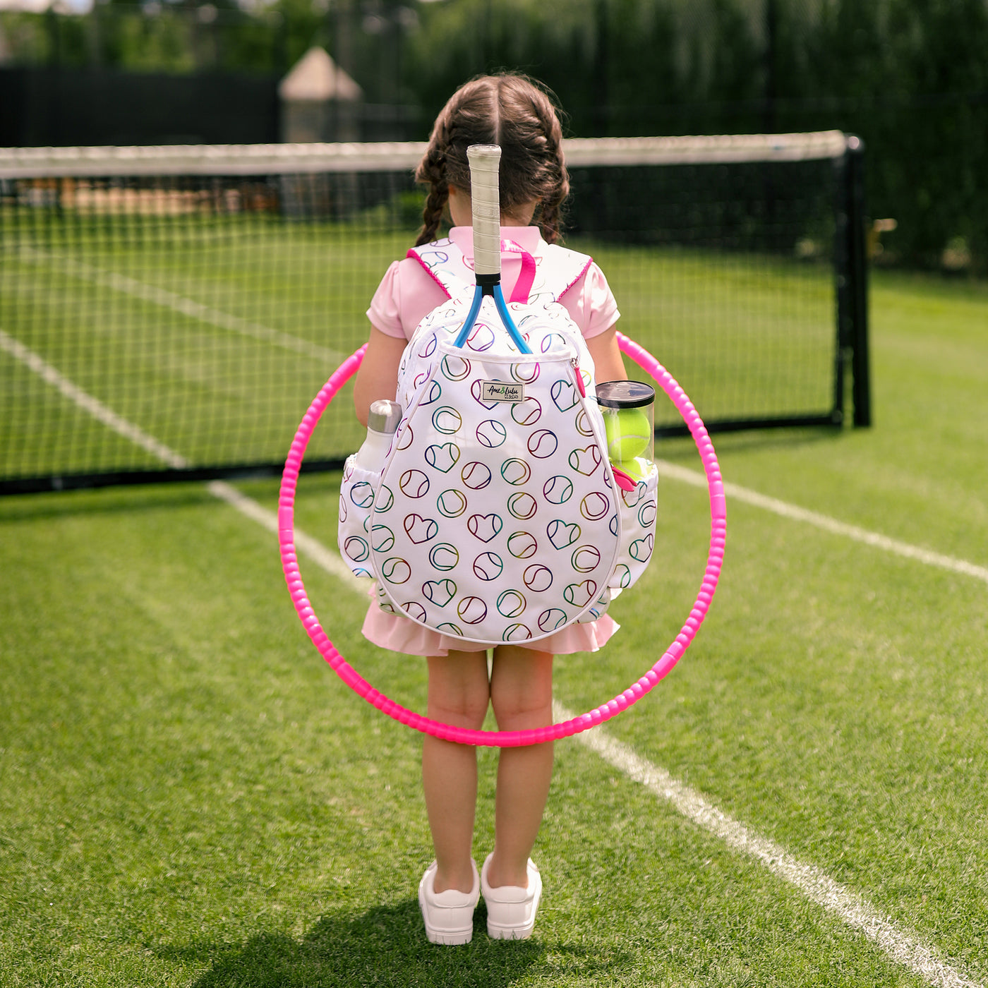 White kids tennis backpack with hot pink zippers and metallic rainbow tennis balls and hearts printed on the fabric.